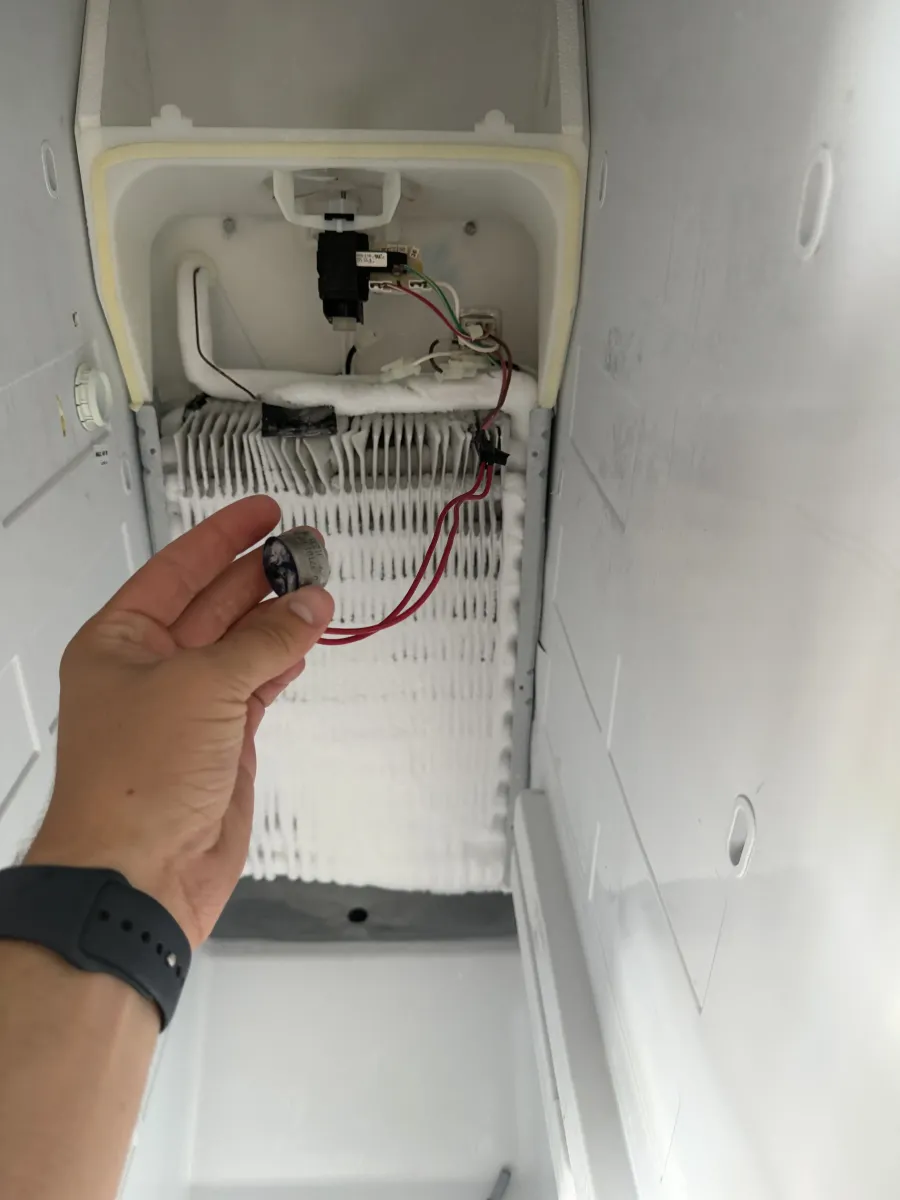 Technician holding a failed defrost thermostat in front of a frosted refrigerator evaporator